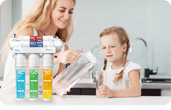 A woman pours water from a pitcher into a glass for a young girl at a kitchen counter. An image of a Brio Legacy water filtration system is overlaid in the foreground.
